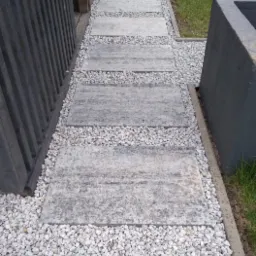 Ścieżka z szarych płyt chodnikowych, otoczona białym grysem, prowadząca wzdłuż ciemnej, drewnianej konstrukcji.