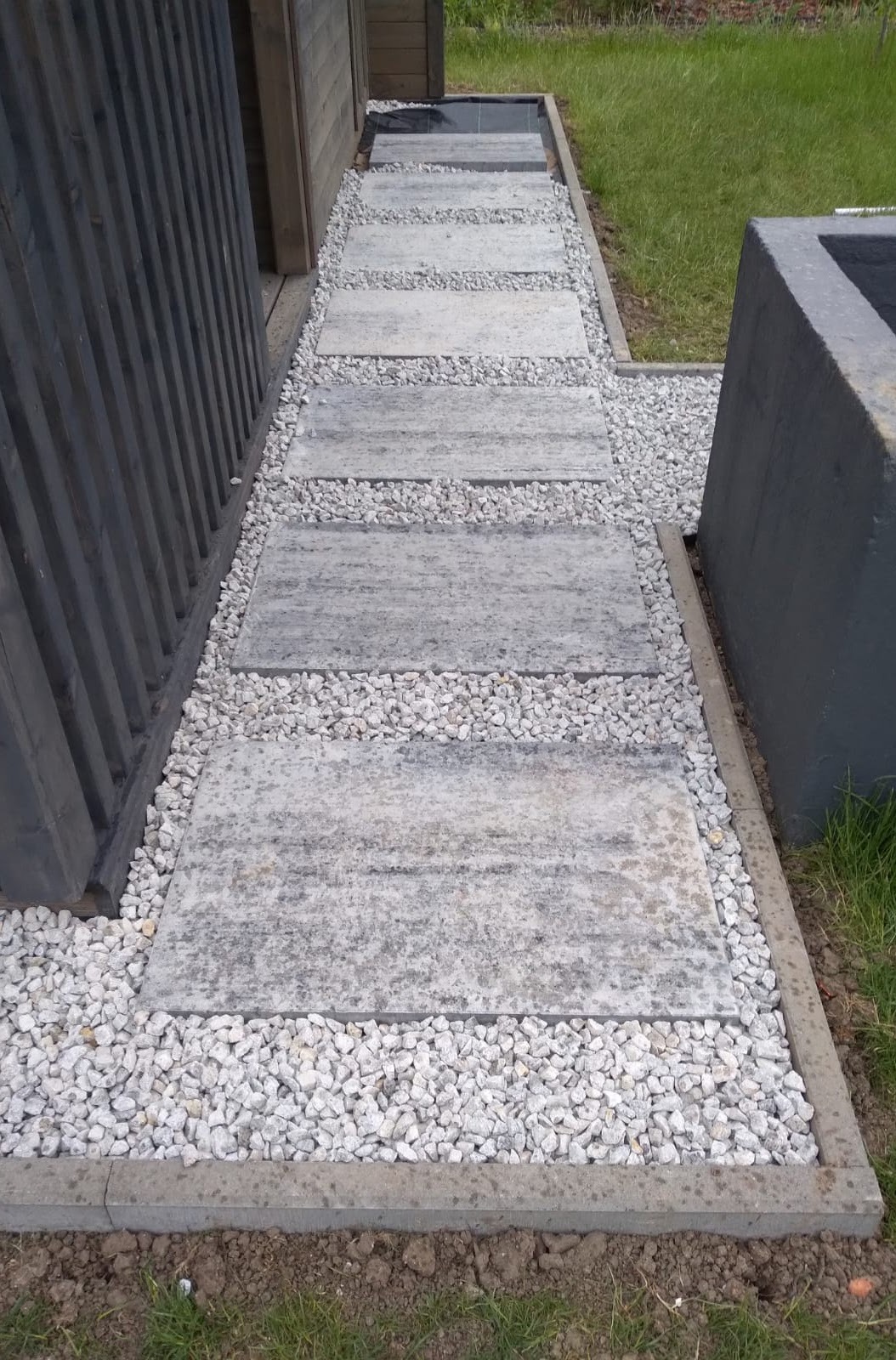 Ścieżka z szarych płyt chodnikowych, otoczona białym grysem, prowadząca wzdłuż ciemnej, drewnianej konstrukcji.