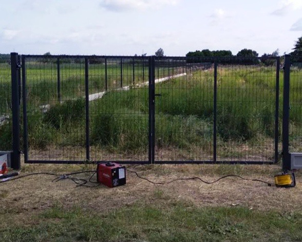 Czarna, metalowa, dwuskrzydłowa brama siatkowa w trakcie montażu na zarośniętej działce, widoczna spawarka i lampa robocza.