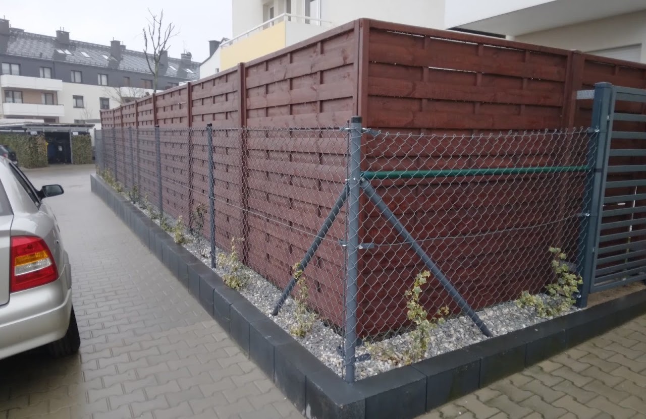 Długi, kombinowany płot: górna część z ciemnobrązowych, poziomych desek, dolna część z metalowej siatki. Wzdłuż płotu rosną młode pnącza, a obok stoi szary samochód osobowy.