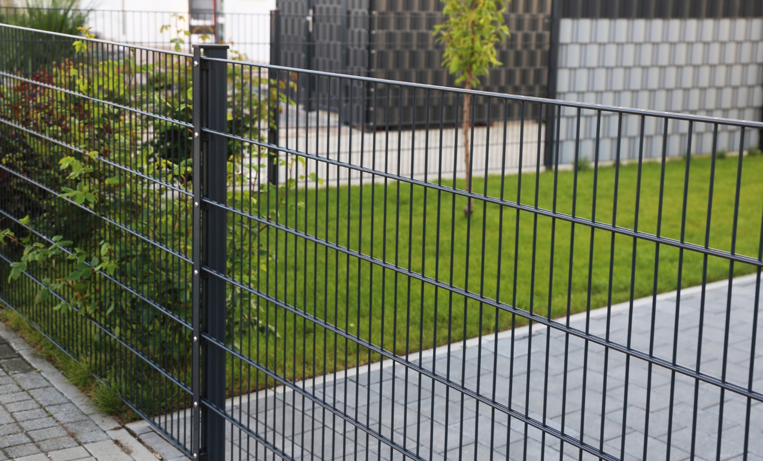 Nowoczesne, antracytowe ogrodzenie panelowe z podwójnego drutu, widok z bliska na słupek i przęsło, z zielonym trawnikiem i krzewami w tle.