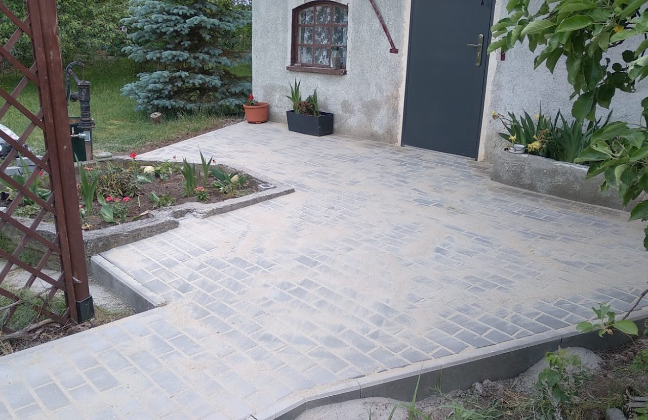 Nowo ułożona szara kostka brukowa w jodełkę, zasypana piaskiem, obok rabatki kwiatowe i wejście do budynku z szarymi drzwiami.