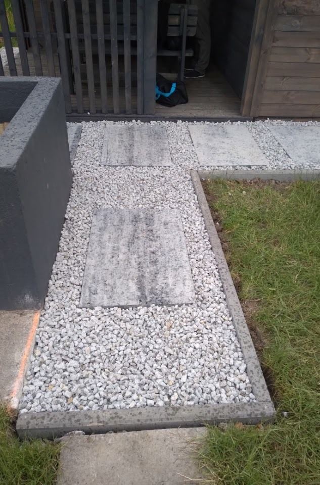 Ścieżka z szarych płyt betonowych oddzielonych białym żwirem prowadząca do drewnianej altany z czarnym ogrodzeniem.