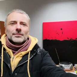 Portret mężczyzny z szarymi włosami w kurtce na tle abstrakcyjnego obrazu w odcieniach czerwieni i czerni oraz biurka z radiem. Nietypowe tło dla grafika.