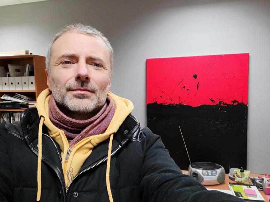 Portret mężczyzny z szarymi włosami w kurtce na tle abstrakcyjnego obrazu w odcieniach czerwieni i czerni oraz biurka z radiem. Nietypowe tło dla grafika.