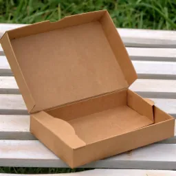 Otwarta, pusta, brązowa tekturowa skrzynka z wieczkiem na białym, pomalowanym drewnianym stole, z zielonym trawiastym tłem.