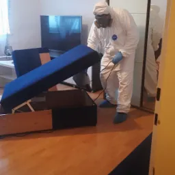 Pracownik w kombinezonie ochronnym i masce opryskuje wnętrze rozkładanej sofy w mieszkaniu, widoczny sprzęt do oprysku.