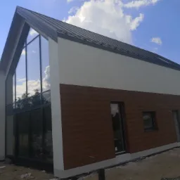 Nowoczesny dom jednorodzinny z dużymi oknami i elewacją w kolorze białym i imitacji drewna, widok z boku na tle błękitnego nieba z chmurami.