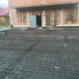 Zbrojenie fundamentu domu jednorodzinnego z widocznym szkieletem budynku w tle i żółtymi rurami instalacyjnymi.