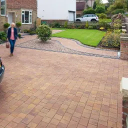 Szeroki podjazd z kostki brukowej w odcieniach brązu i beżu, z dekoracyjną ścieżką z kamyków i zadbanym trawnikiem, kobieta z torbą idzie w stronę domu z cegły.