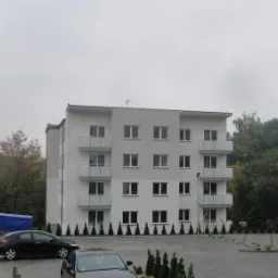 Nowoczesny, czterokondygnacyjny budynek mieszkalny z balkonami, otoczony drzewami i zaparkowanymi samochodami, sfotografowany w pochmurny dzień.