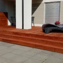 Nowoczesny taras z drewna egzotycznego w Pęgowie, z trzema stopniami i czarnym leżakiem z poduszką, na tle białego domu z szarymi roletami.