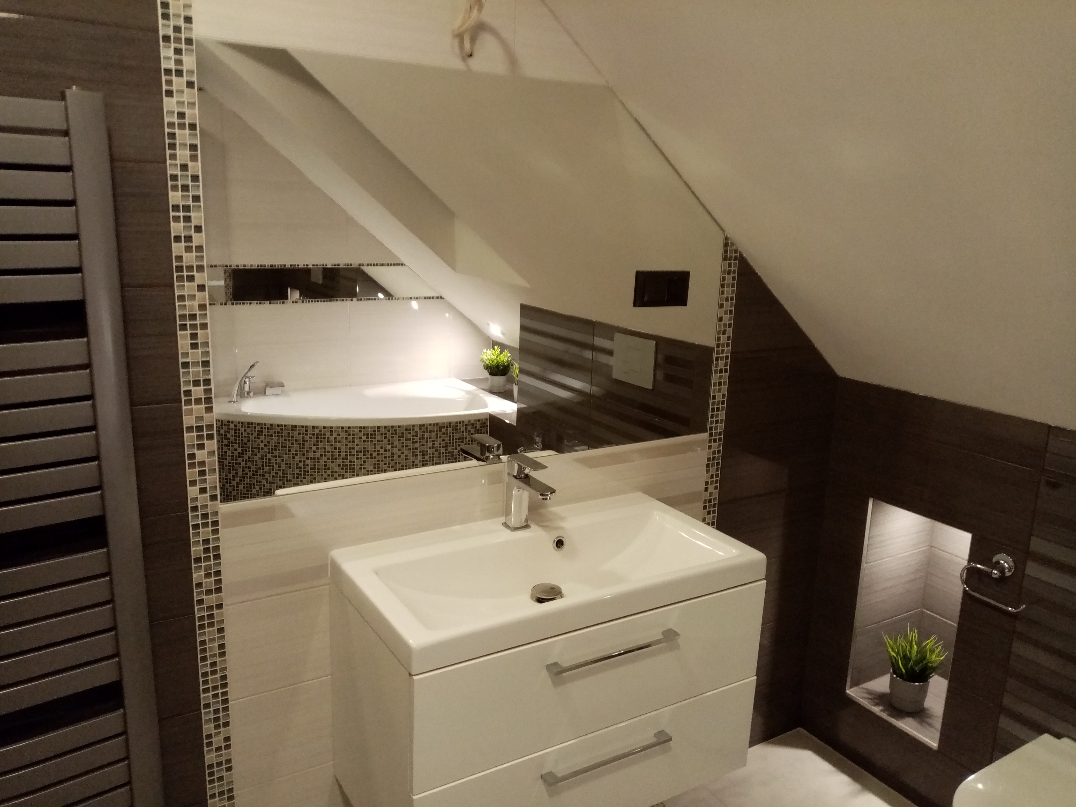 Nowoczesna łazienka na poddaszu z umywalką na szafce, grzejnikiem drabinkowym, lustrem i wanną narożną w tle, wykończona mozaiką i płytkami imitującymi drewno.