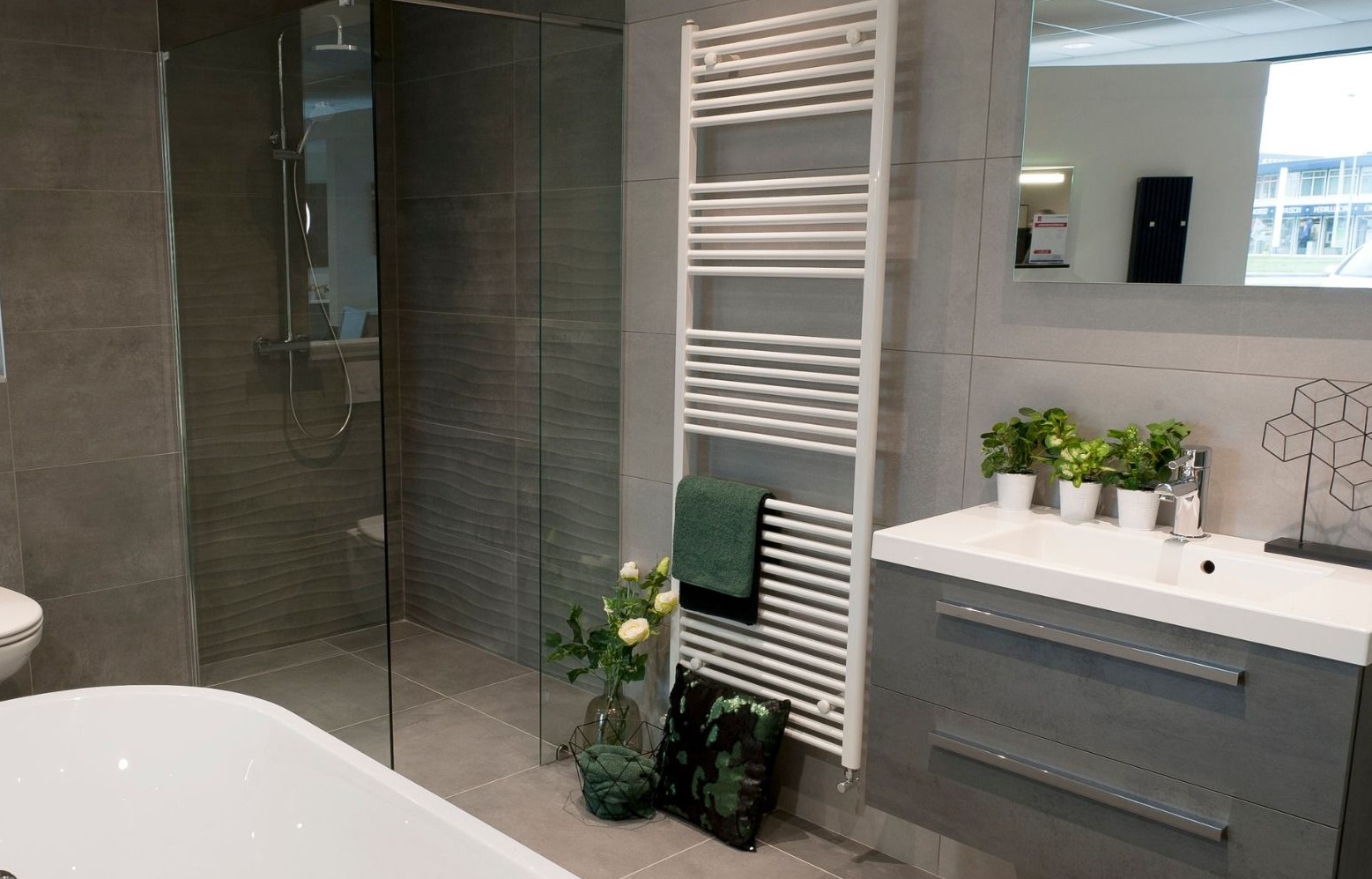 Szara łazienka z kabiną prysznicową, grzejnikiem drabinkowym z zielonym ręcznikiem, umywalką z szafką i dekoracjami roślinnymi. W tle widoczne odbicie w lustrze.