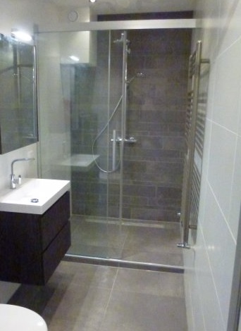 Nowoczesna łazienka z kabiną prysznicową typu walk-in, ciemnymi płytkami imitującymi cegłę na ścianie prysznica, białą umywalką nablatową na ciemnobrązowej szafce i grzejnikiem drabinkowym.