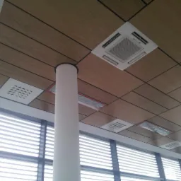 Kasetonowy sufit z drewnopodobnymi panelami, wbudowanym klimatyzatorem Mitsubishi i kratkami wentylacyjnymi, widok z dołu na kolumnę i żaluzje okienne.