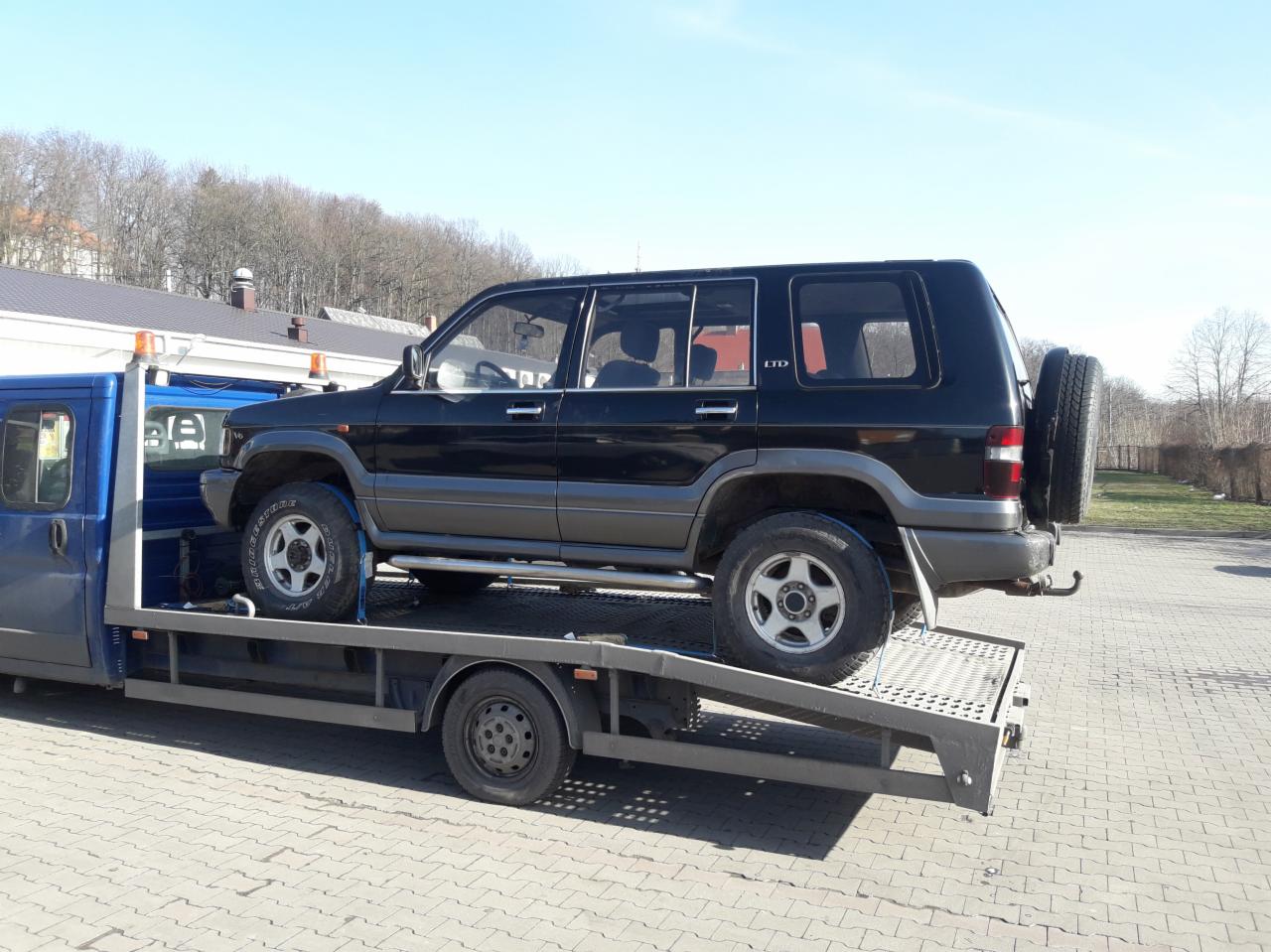 Czarny samochód terenowy marki Isuzu na platformie lawety w słoneczny dzień.