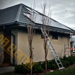 Dom jednorodzinny z panelami fotowoltaicznymi na dachu, drabiny opierają się o ścianę, pochmurne niebo w tle.