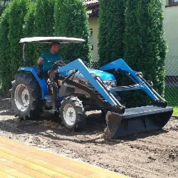 Operator na niebieskim traktorze z ładowarką wyrównuje teren przygotowany pod trawnik, widoczne świeże ślady opon i fragment drewnianego tarasu.
