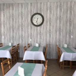 Wnętrze restauracji z szarą tapetą imitującą drewniane panele, zegarem na ścianie i nakrytymi stołami z drewnianymi krzesłami.