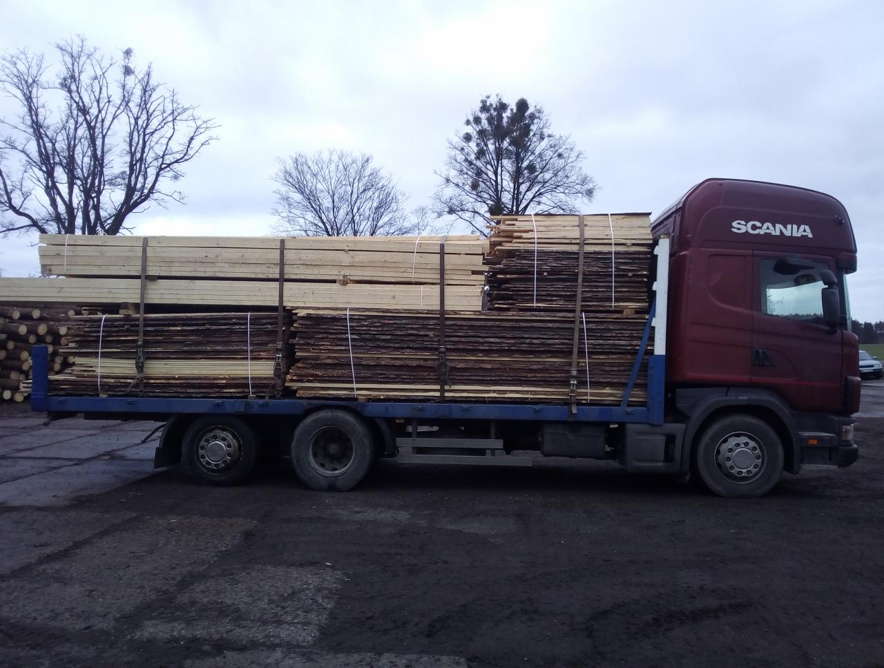 Ciężarówka Scania z niebieską platformą załadowana drewnem, zabezpieczonym pasami, na tle pochmurnego nieba i drzew bez liści.