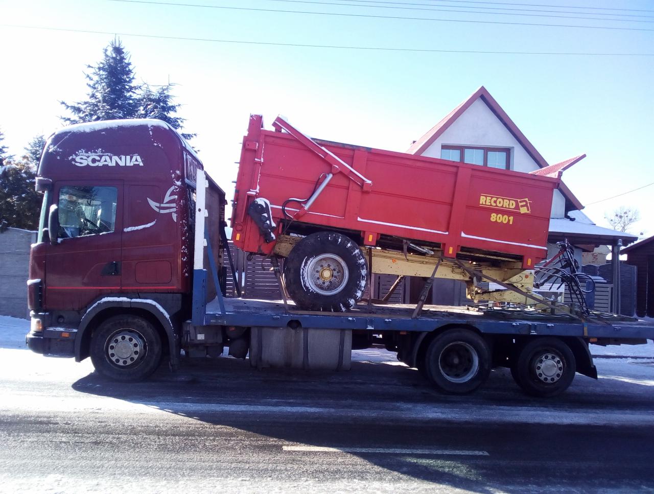 Ciężarówka Scania z czerwoną przyczepą rolniczą Record 8001 na platformie, zimowy krajobraz w tle.