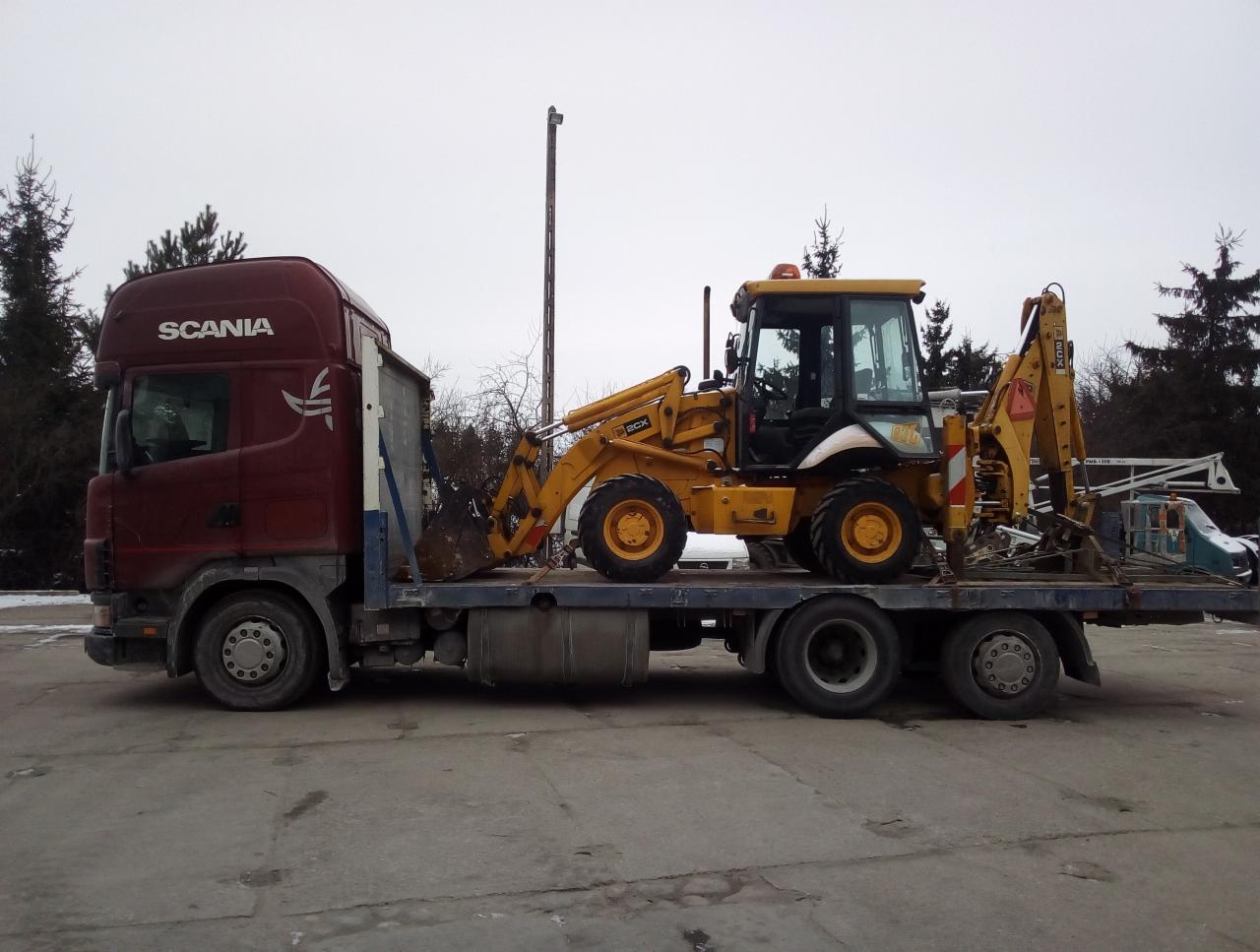 Czerwony ciągnik siodłowy marki Scania z naczepą niskopodwoziową przewożący żółtą koparko-ładowarkę JCB 2CX na tle pochmurnego nieba i drzew iglastych.