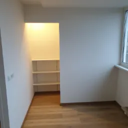 Wnętrze pokoju po remoncie: świeżo pomalowane białe ściany, drewnopodobna podłoga, wnęka z półkami oświetlona od góry, listwa przypodłogowa w kolorze drewna.