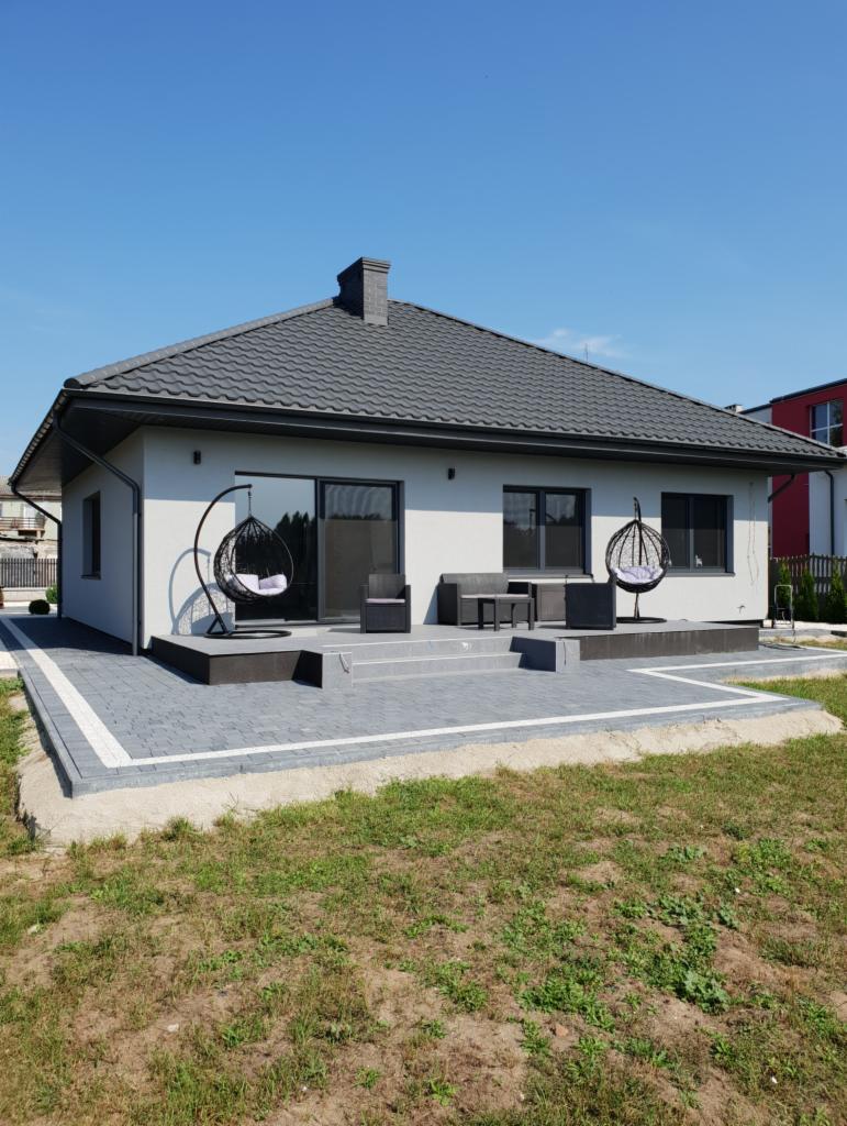 Nowoczesny, parterowy dom z szarą dachówką i tarasem wyłożonym kostką brukową. Na tarasie wiszące fotele typu kokon i zestaw wypoczynkowy.