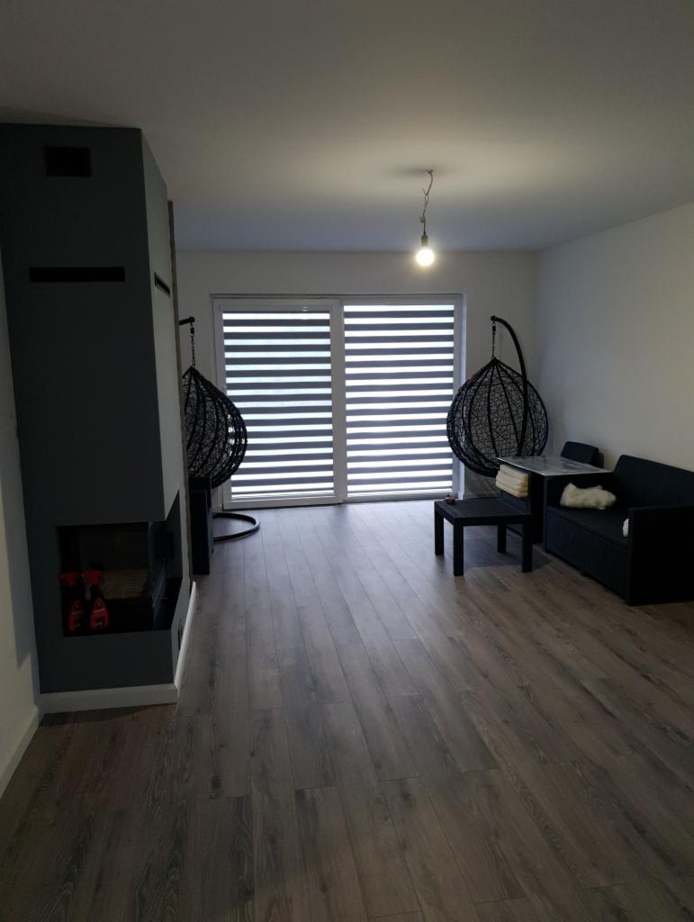 Nowoczesny salon z ciemną podłogą, szarym kominkiem, wiszącymi fotelami i przesuwnymi drzwiami z żaluzjami poziomymi.