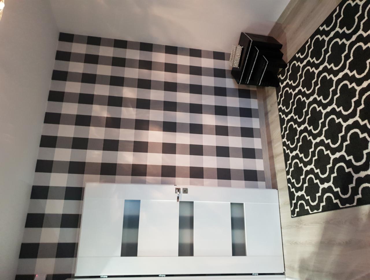 Ściana w czarno-białą kratę nad białymi drzwiami, obok czarny dywan w białe wzory i czarna szafka nocna z kryształkami.