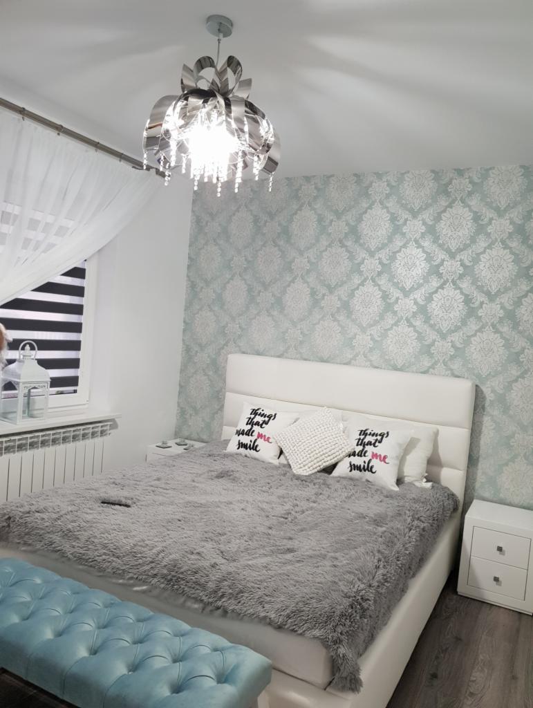 Nowoczesna sypialnia z tapetą w delikatne wzory, łóżkiem z pluszową narzutą, ozdobnymi poduszkami i srebrnym żyrandolem.