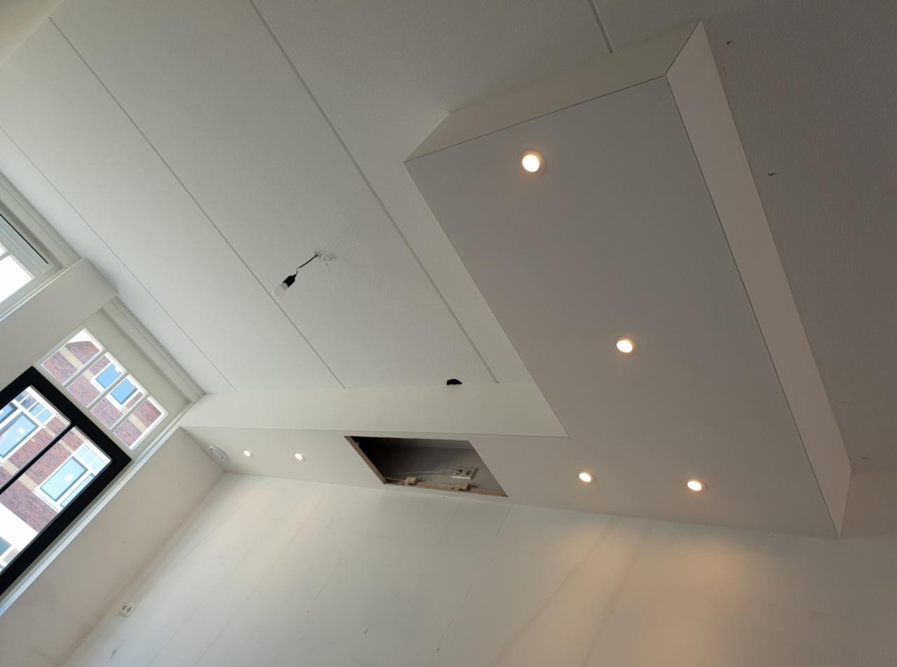 Nowoczesny biały sufit z wbudowanymi punktami świetlnymi, widoczne niedokończone miejsce po montażu oświetlenia i wisząca żarówka na kablu, okno z widokiem na budynek z cegły.