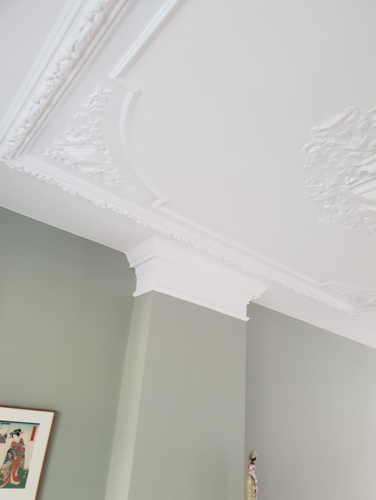Biały sufit z dekoracyjnymi sztukateriami i listwami przyściennymi, widok z dołu na szaro-zielone ściany z obrazem i figurką.