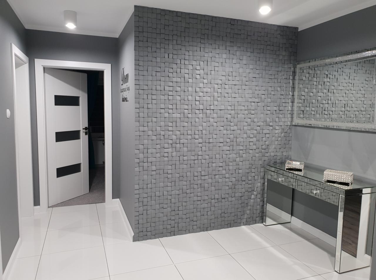 Szara ściana z trójwymiarowych kwadratów, lustrzana konsola z dekoracyjnymi szkatułkami, biała podłoga i drzwi w nowoczesnym przedpokoju.