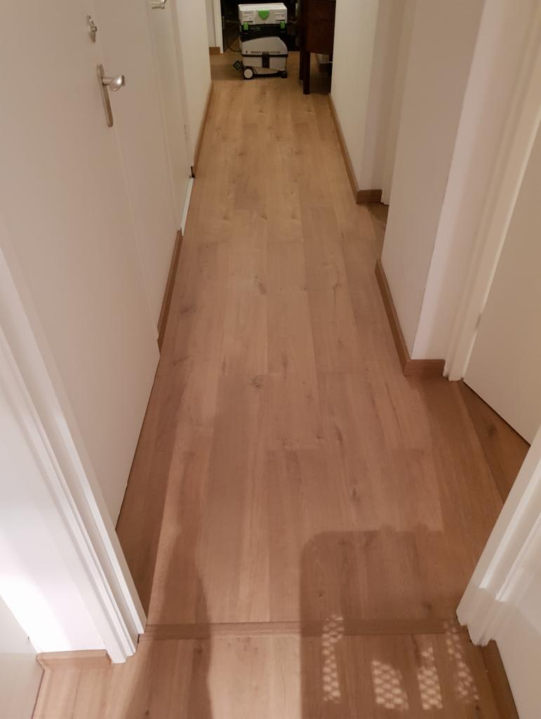 Korytarz z nową, jasną drewnianą podłogą, widoczny odkurzacz przemysłowy w tle.