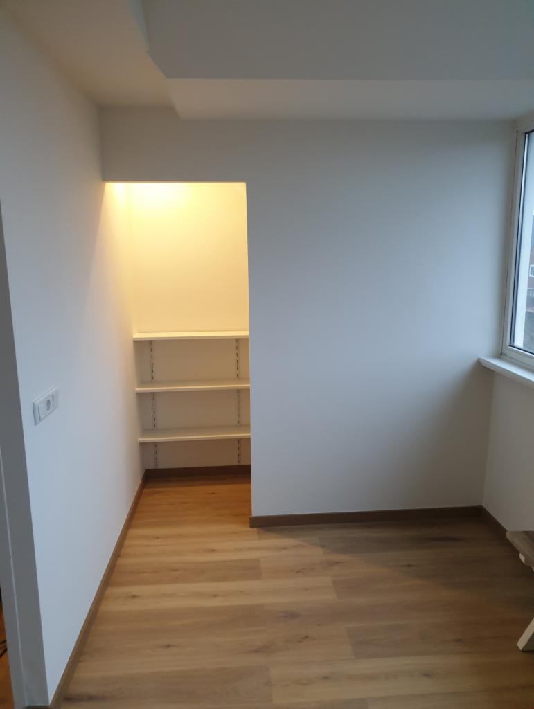 Wnętrze pokoju po remoncie: świeżo pomalowane białe ściany, drewnopodobna podłoga, wnęka z półkami oświetlona od góry, listwa przypodłogowa w kolorze drewna.
