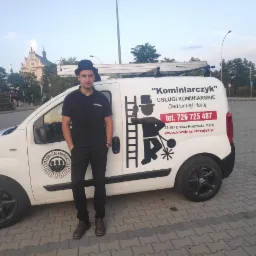 Kominiarz w czarnym stroju i meloniku stoi przed białym samochodem firmowym z logo i danymi kontaktowymi na tle budynku kościoła