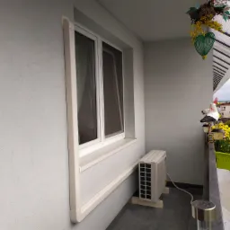 Biała jednostka zewnętrzna klimatyzatora zamontowana na balkonie, widok na okno z ramą i ozdobne dekoracje balkonowe.