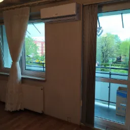 Wnętrze pokoju z zamontowaną jednostką klimatyzacji Gree nad oknem, widok na balkon z kartonami po klimatyzatorze i zielenią w tle.