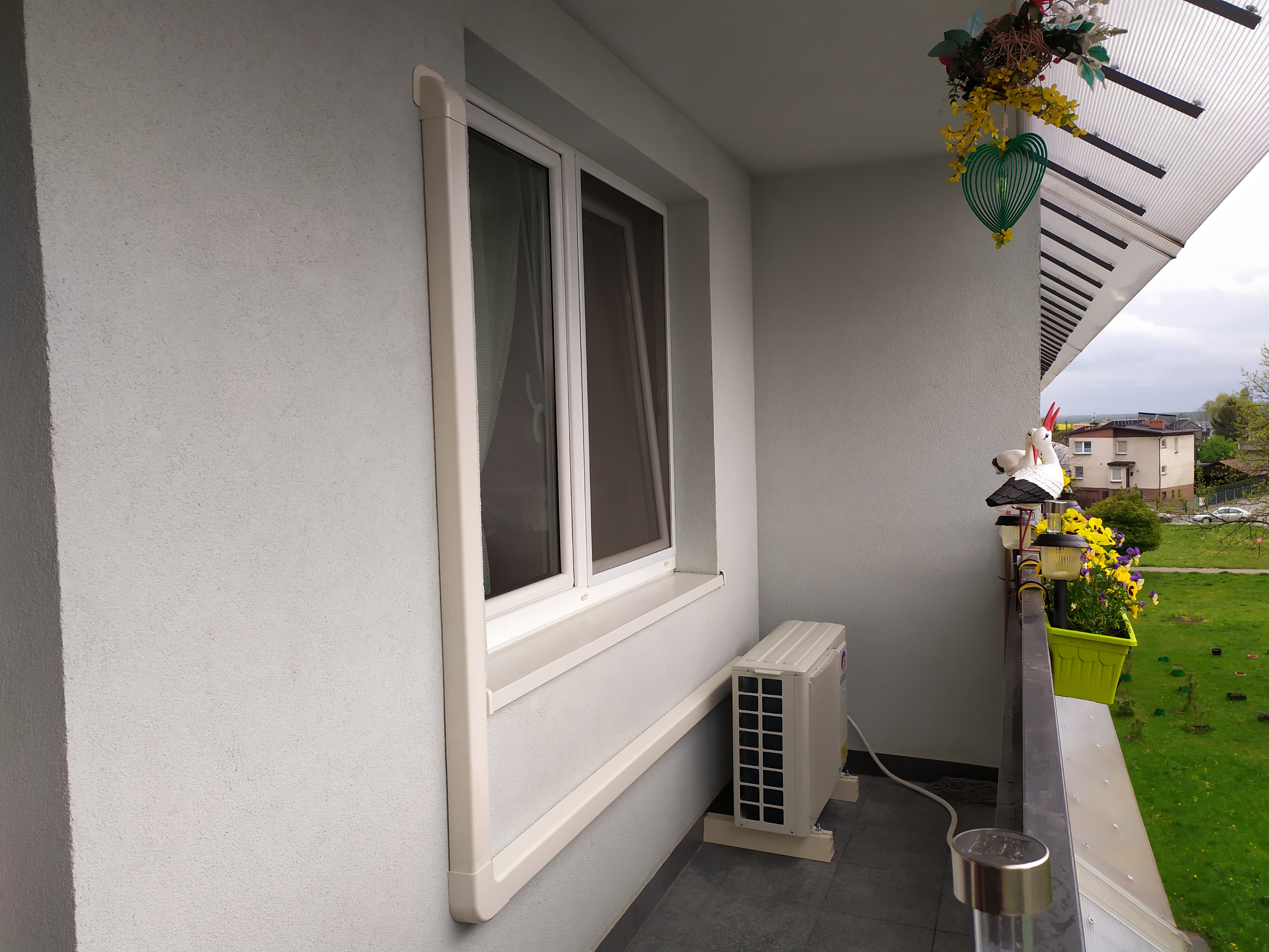 Biała jednostka zewnętrzna klimatyzatora zamontowana na balkonie, widok na okno z ramą i ozdobne dekoracje balkonowe.