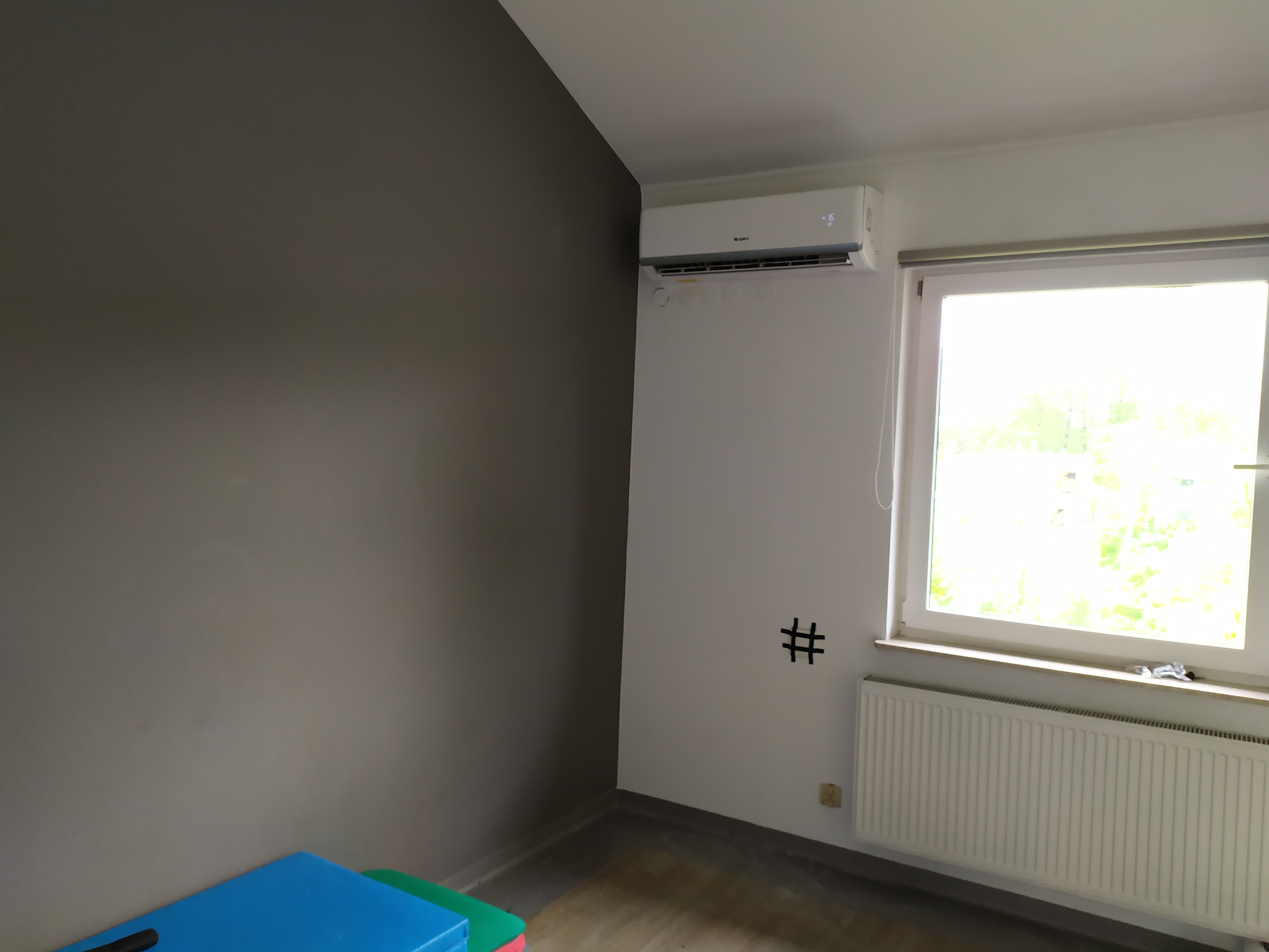 Biały klimatyzator na ścianie w pokoju z szarą ścianą, widoczne ślady po instalacji, grzejnik pod oknem.