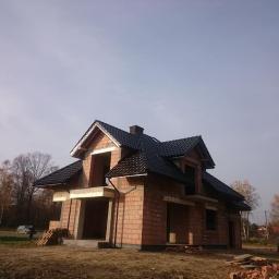 TMK DACH - Dom w trakcie budowy z częściowo ukończonym dachem z czarnej dachówki, widoczne elementy konstrukcyjne i cegły.