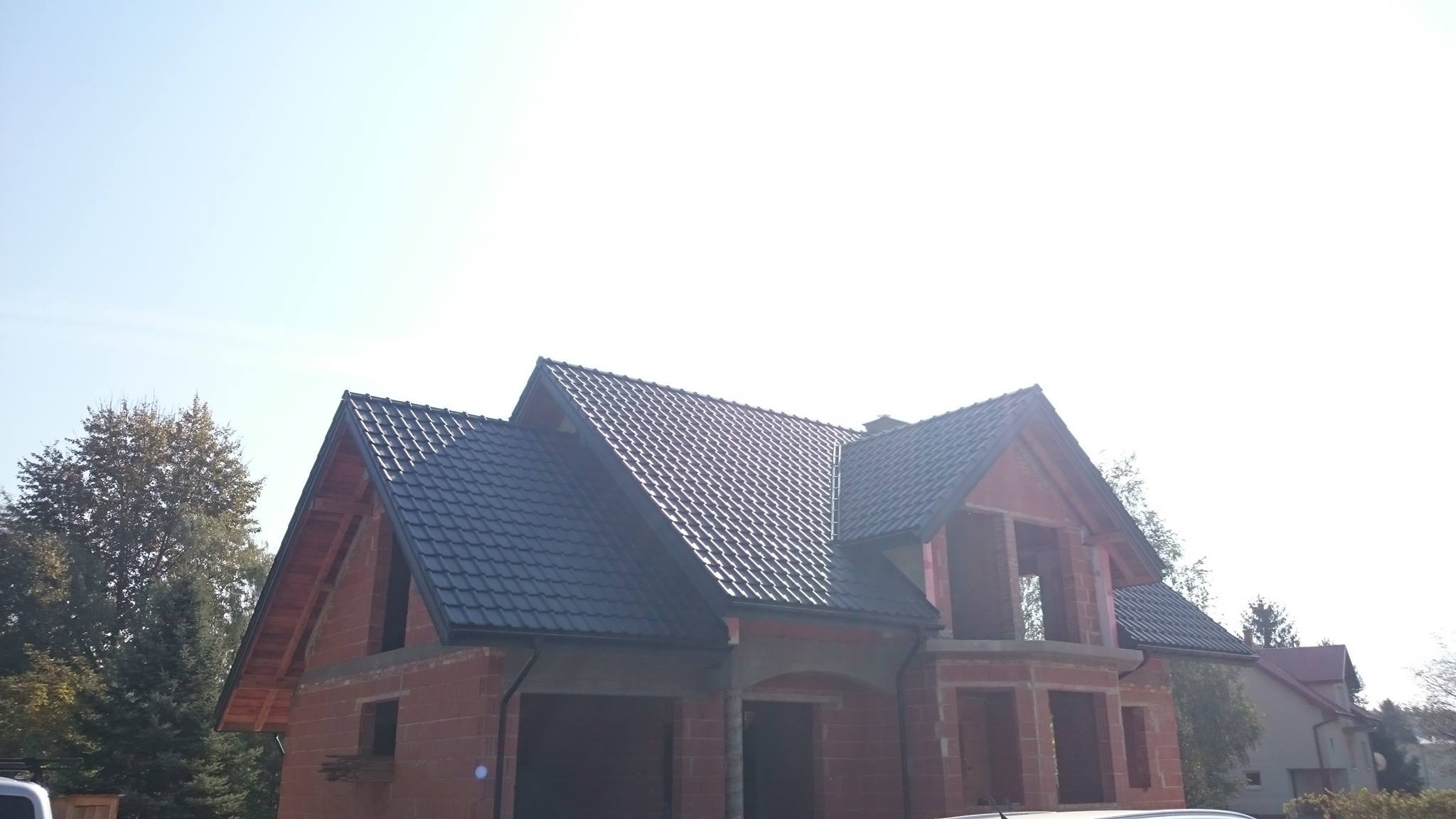 Nowy, ciemnoszary dach z dachówki ceramicznej na surowym budynku z czerwonej cegły, widoczne elementy więźby dachowej, jasne niebo w tle.