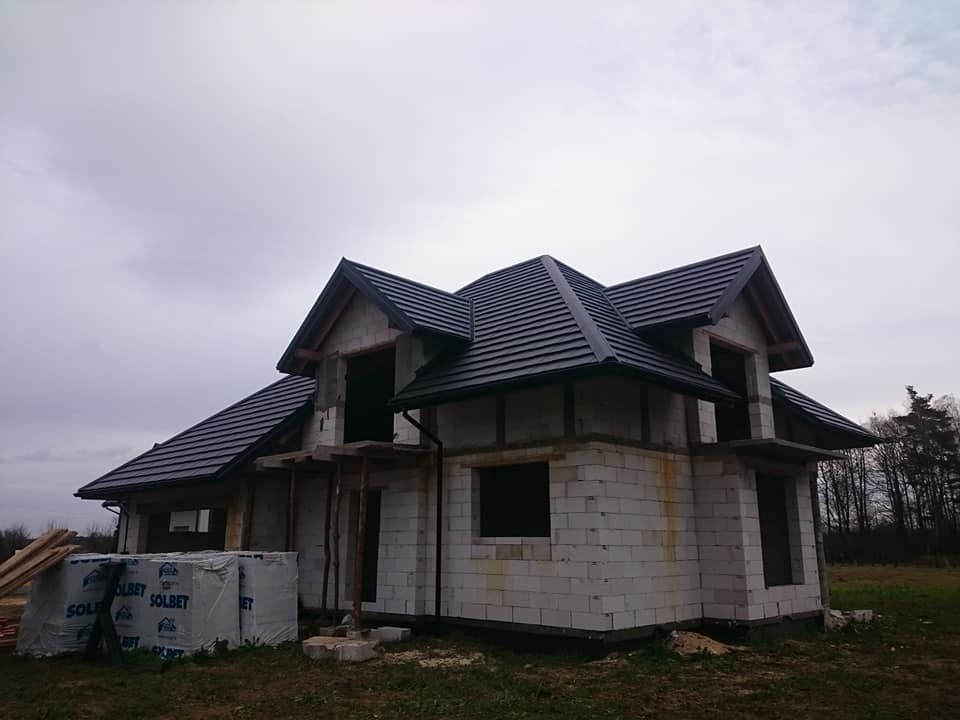 Dom w trakcie budowy z częściowo ukończonym, czarnym dachem z dachówki ceramicznej, widoczne materiały budowlane Solbet i elementy konstrukcji drewnianej dachu.