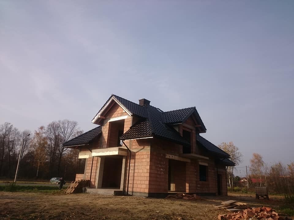 Dom w trakcie budowy z częściowo ukończonym dachem z czarnej dachówki, widoczne elementy konstrukcyjne i cegły.