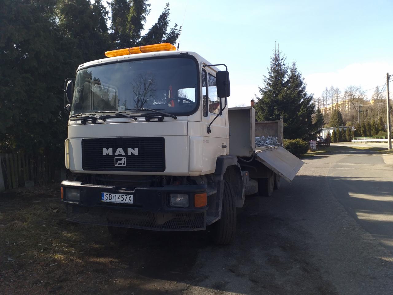 Biała ciężarówka MAN z pomarańczowym światłem ostrzegawczym na dachu, platforma załadunkowa częściowo opuszczona, widoczny ładunek kamieni.