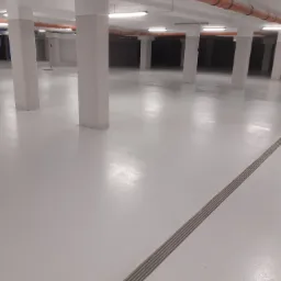 Biała, gładka posadzka żywiczna w hali magazynowej z widocznym odpływem liniowym i pomarańczowymi rurami pod sufitem.