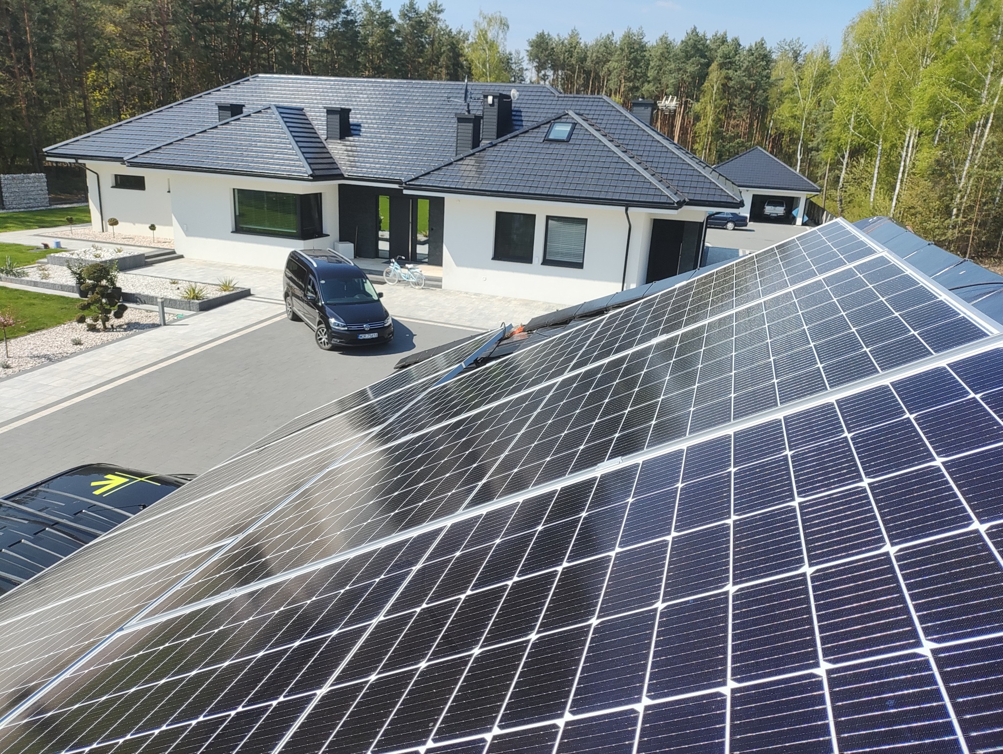 Nowoczesny dom z panelami fotowoltaicznymi na dachu, widok z góry. Na podjeździe stoi czarny van. W tle drzewa i błękitne niebo.