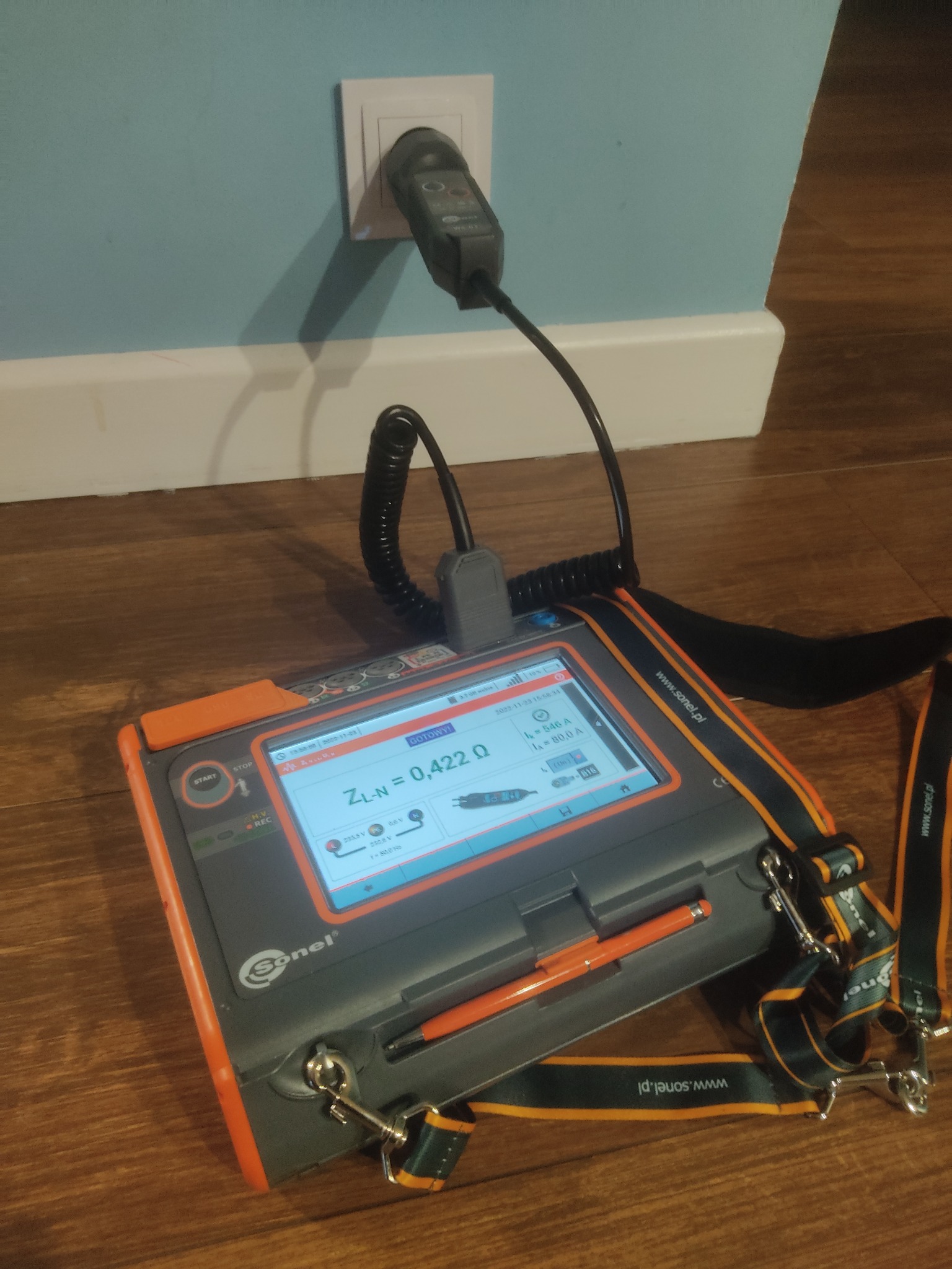 Miernik Sonel podłączony do gniazdka elektrycznego, wyświetlający wynik pomiaru impedancji pętli zwarcia ZL-N = 0,422 Ω, na tle drewnianej podłogi i niebieskiej ściany.